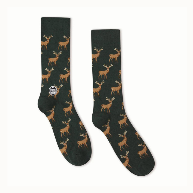 Knitted crew socks by The Landmark Project featuring a repeating pattern of brown pixelated stags with large antlers on a dark forest green background and a small white circular tram logo near the ankle