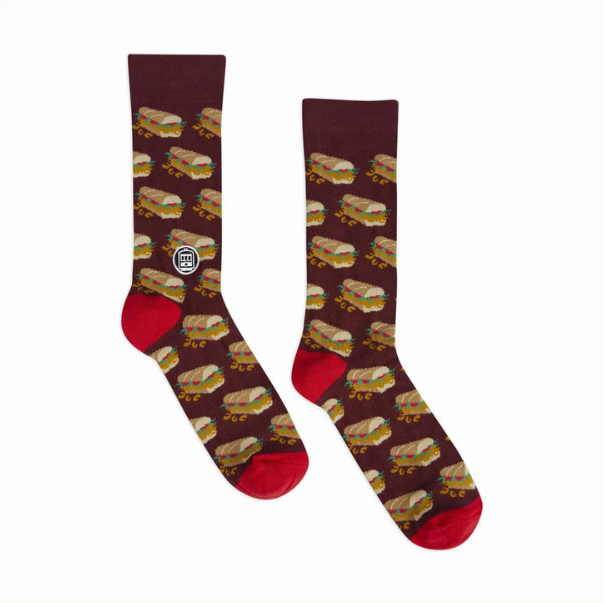Woven crew socks featuring a repeating pattern of New Orleans dressed po-boy sandwiches against a maroon background with a white St. Charles streetcar logo and vibrant red heel and toe accents