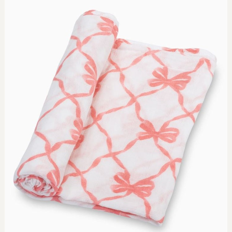 Muslin cotton swaddle blanket by Petite Plume featuring a coral-pink bow-and-ribbon lattice pattern on a white studio background with soft breathable gauze and a partially rolled edge