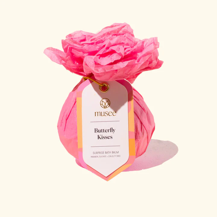 Commercial studio photography for Musee featuring a Butterfly Kisses bath balm wrapped in vibrant pink crinkled tissue paper with a ruffled floral top and a white branded tag secured by a gold ring against a minimalist cream background