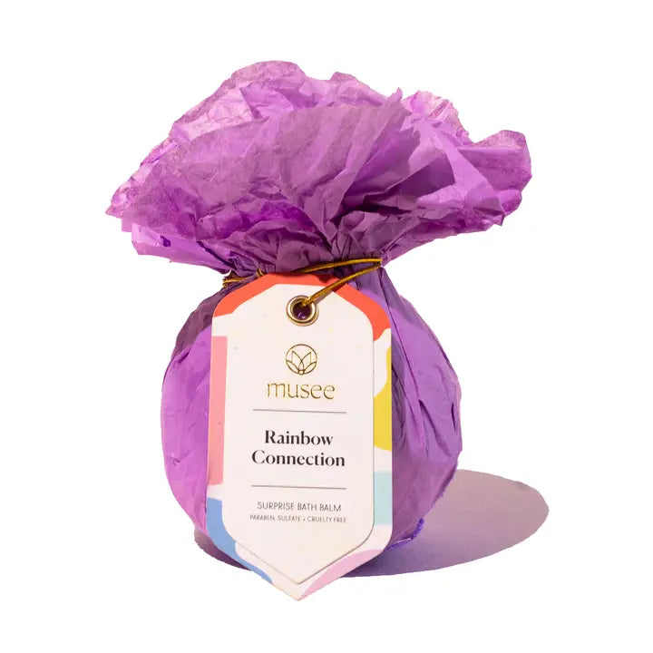 Product photography of a Musee Rainbow Connection bath balm wrapped in vibrant purple crinkled tissue paper featuring a white gift tag with colorful pastel borders and a thin gold elastic cord against a plain white background
