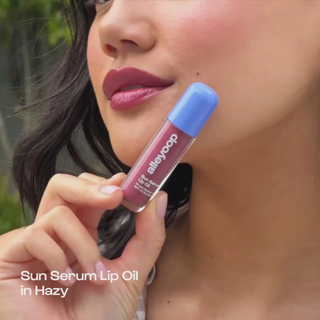 Digital photograph by Alleyoop featuring a woman holding the Sun Serum Lip Oil in Hazy with a periwinkle blue cap, highlighting glossy plum-colored lips and soft pink manicured nails against a natural outdoor background