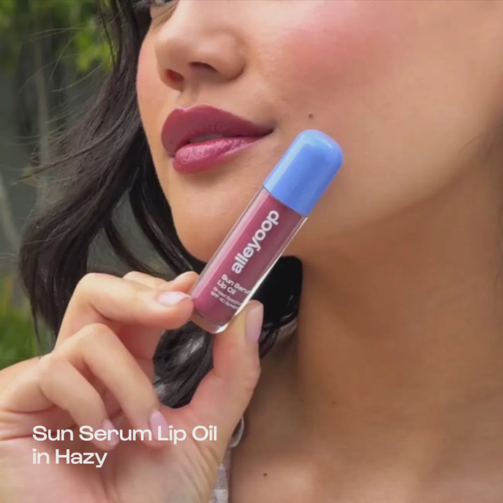 Digital photograph by Alleyoop featuring a woman holding the Sun Serum Lip Oil in Hazy with a periwinkle blue cap, highlighting glossy plum-colored lips and soft pink manicured nails against a natural outdoor background