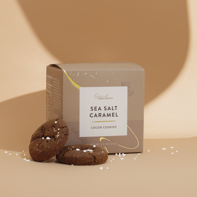 Studio product photography for Grand Provisions featuring a minimalist tan box of Sea Salt Caramel Cocoa Cookies next to two dark cookies topped with coarse white salt flakes on a warm beige background with soft curved shadows