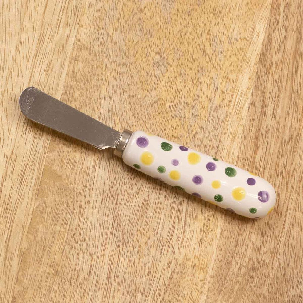 Photograph of a stainless steel butter spreader on a light wood grain surface featuring a decorative white ceramic handle adorned with vibrant yellow, purple, and green polka dots