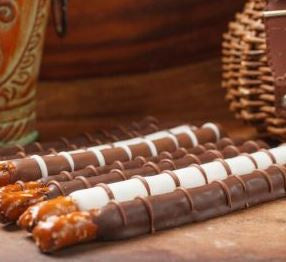 Photograph of chocolate-covered pretzel rods on a wooden surface featuring alternating patterns of white and milk chocolate drizzle with visible salt crystals against a rustic woven basket background
