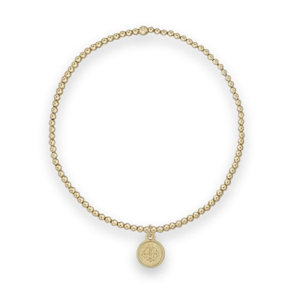 Gold-tone beaded stretch bracelet featuring small polished spherical beads, a single larger accent bead, and an iconic St. Benedict religious medallion pendant centered on a light grey background