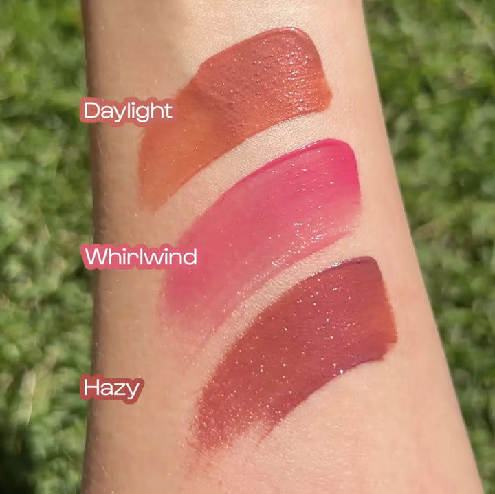 Photograph of three glossy makeup swatches on a forearm outdoors against a green lawn featuring a terracotta orange shade labeled Daylight a vibrant pink named Whirlwind and a deep rose called Hazy with shimmering light-reflecting particles