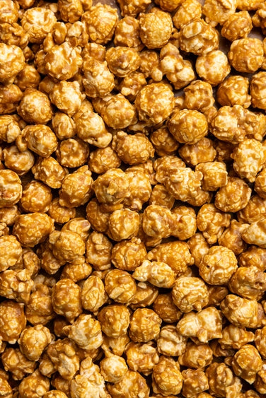 Digital photography of a dense pile of caramel popcorn featuring glossy golden-brown glazed kernels and a crunchy sugar-coated texture in an overhead macro view