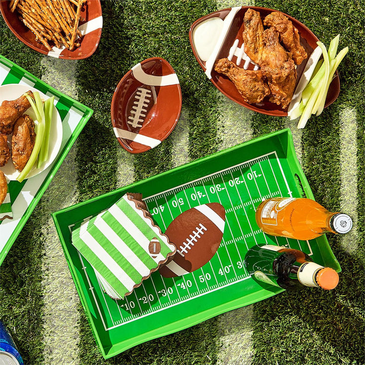 Digital photograph of a football-themed snack spread on artificial turf featuring a green field-design tray with white yard line markings, brown football-shaped ceramic bowls filled with crispy wings, and green and white striped napkins with a football icon