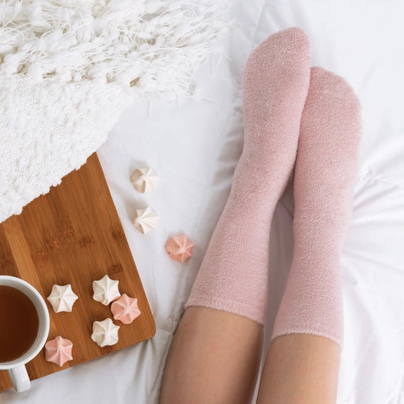 Photograph of a person wearing soft pink fuzzy socks on a white bed next to a wooden tray featuring pastel star-shaped meringues and a textured white knit blanket