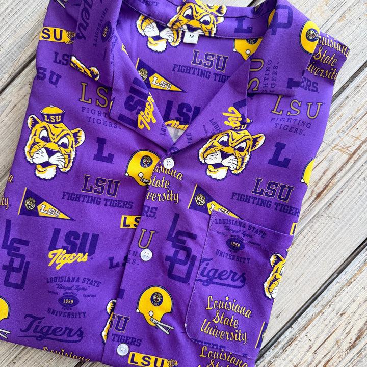 Cotton apparel by LSU brand featuring a purple button-down shirt folded on a rustic wooden background with yellow Mike the Tiger mascot logos, gold Fighting Tigers typography, and white buttons