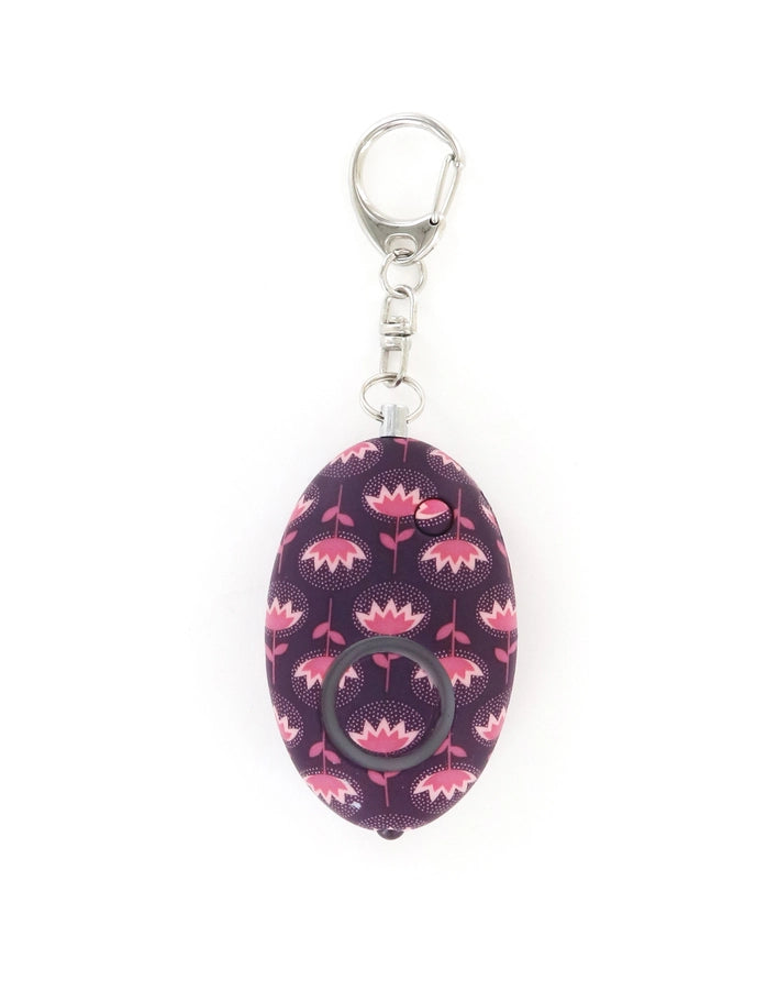 Product photograph of a personal safety alarm keychain featuring an oval purple body with stylized pink flowers and white pointillist halos attached to a silver metal lobster clasp against a white background
