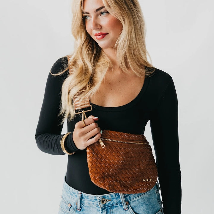 Studio photograph of a blonde woman modeling a brown woven leather crossbody bag with a wide striped strap, gold jewelry, and a black long-sleeved bodysuit against a white studio background