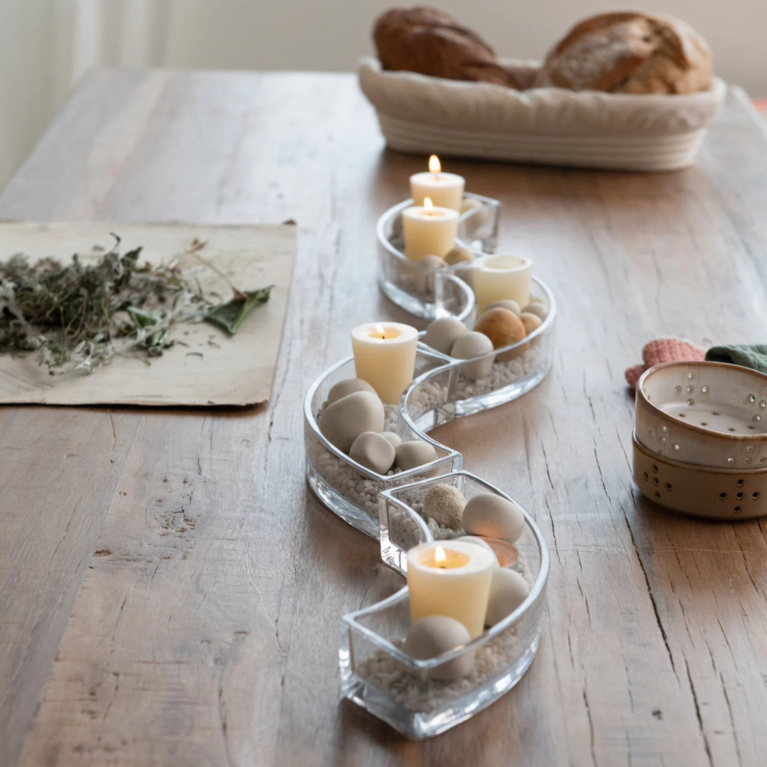 A high-angle photograph of a serpentine clear glass candle holder on a rustic wooden dining table featuring lit cream pillar candles nestled in smooth grey pebbles and a basket of crusty artisan bread in the background
