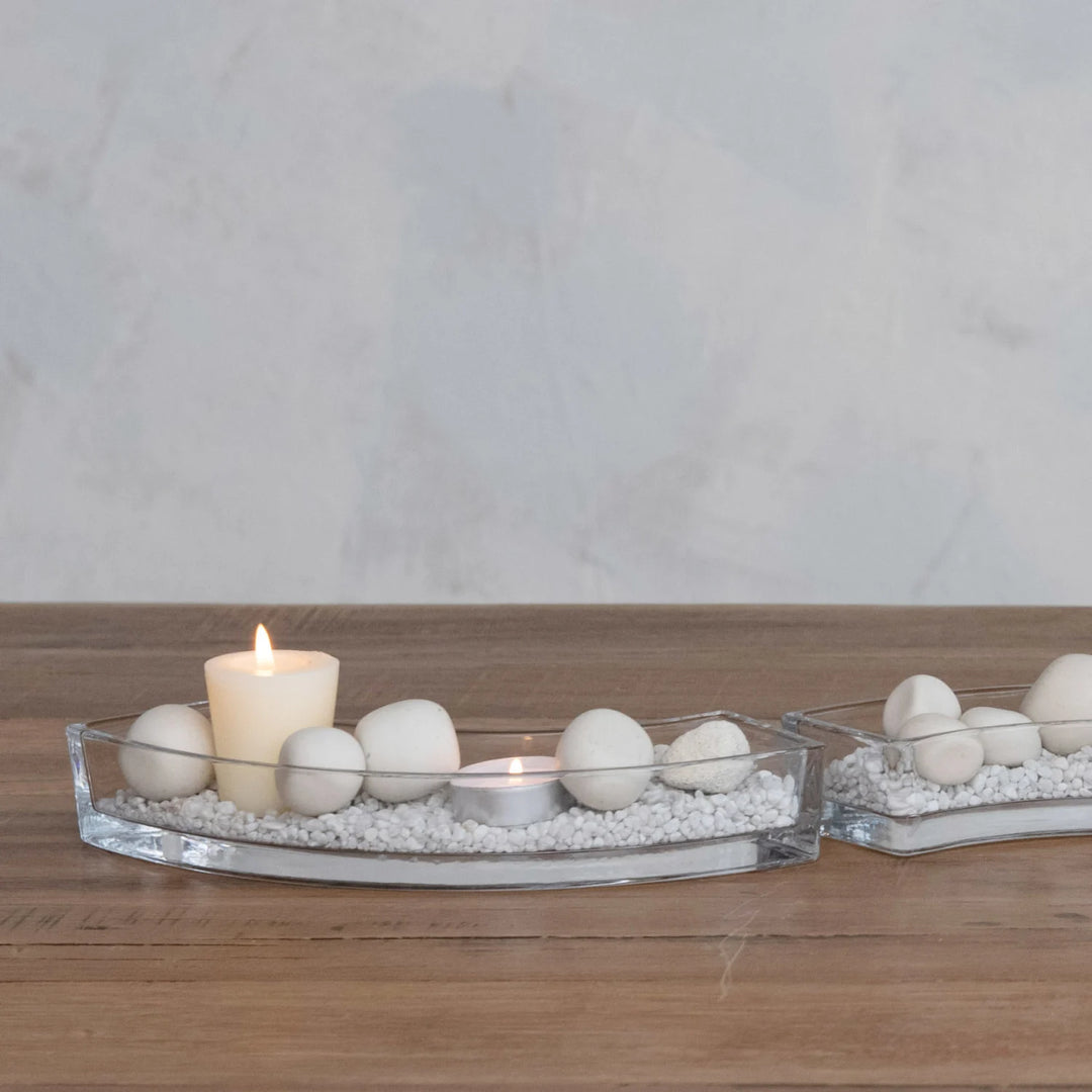 Photograph of minimalist curved glass trays on a wooden tabletop featuring smooth white ceramic spheres and pebbles arranged around a lit pillar candle and a flickering tealight against a textured neutral wall