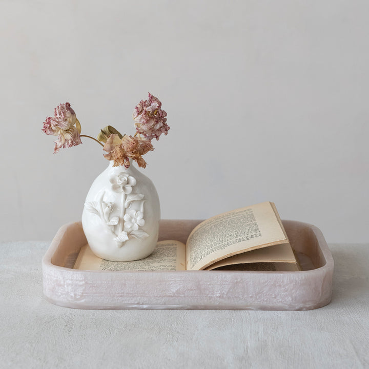 Photograph of a minimalist still life on a neutral tabletop featuring a white ceramic vase with embossed floral relief holding dried pink carnations and an open vintage book resting inside a marble-textured pale pink rectangular tray