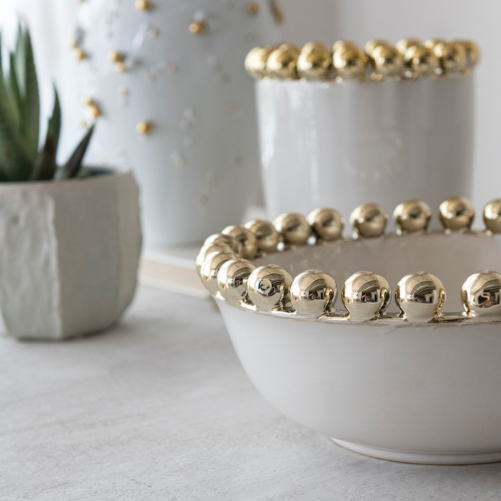 Handcrafted ceramic bowl and vessels by L'Objet on a bright interior tabletop featuring a rim of reflective gold-plated spheres beside a green succulent in a textured gray pot