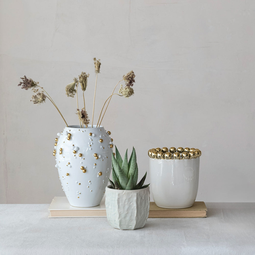 Ceramic pottery by L'Objet featuring a white vase with gold-plated studs and dried flowers alongside a faceted succulent planter and a gold-beaded vessel resting on vintage books in a minimalist setting