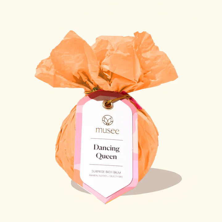 Product photography by Musee of a Dancing Queen surprise bath balm wrapped in peach crinkled tissue paper featuring a pink-accented tag and metallic gold tie on a cream background