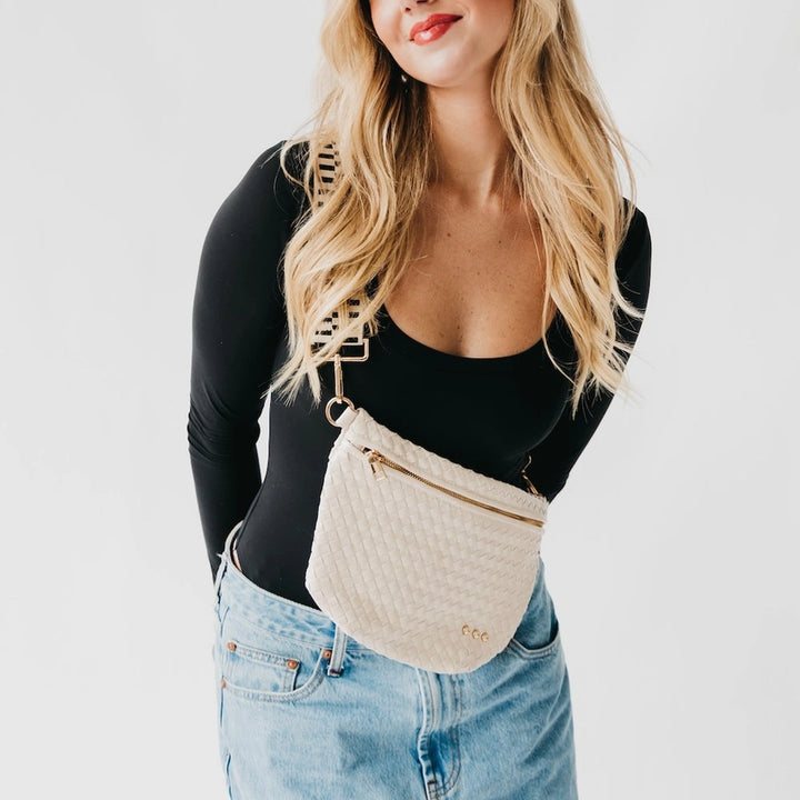 Fashion photograph of a blonde woman with red lipstick wearing a cream woven crossbody bag featuring gold hardware and a geometric patterned strap against a white studio background