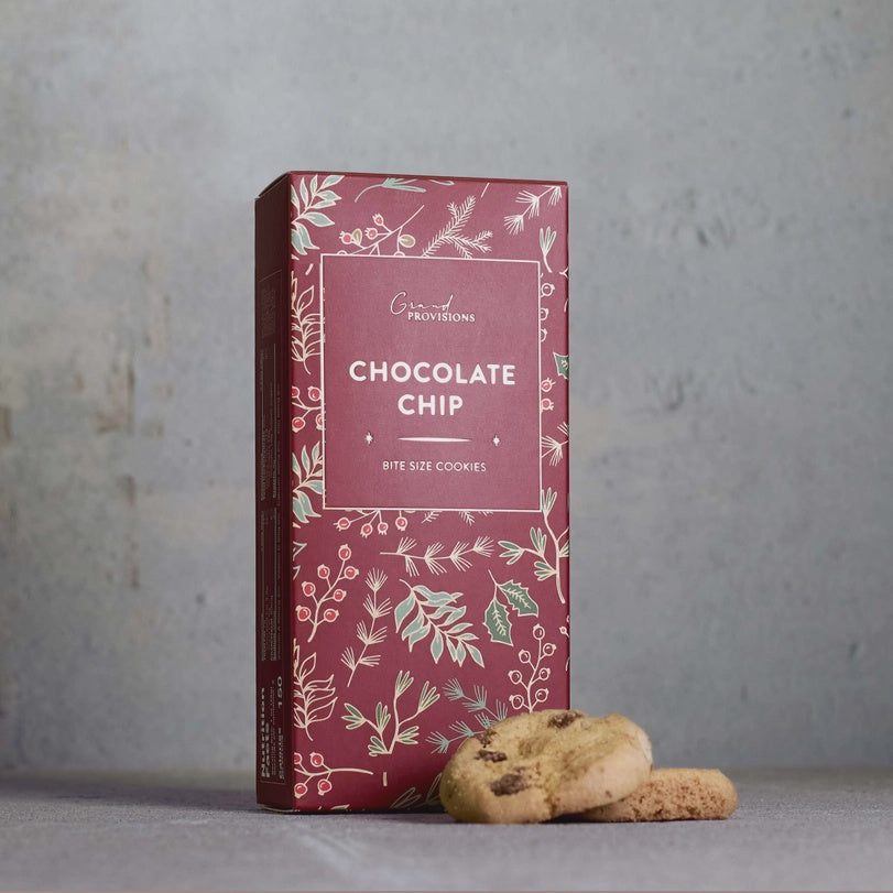 Studio photograph of Grand Provisions chocolate chip bite size cookies featuring a burgundy box with white botanical illustrations and two cookies resting on a textured grey surface