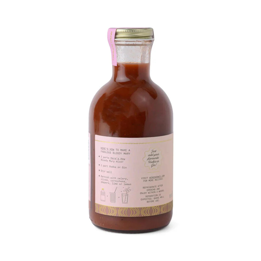 Studio photograph of a Here's How brand Bloody Mary mixer bottle on a white background showcasing the thick red pulpy liquid, a light pink label with decorative gold wave patterns, and a pink security seal across the silver cap