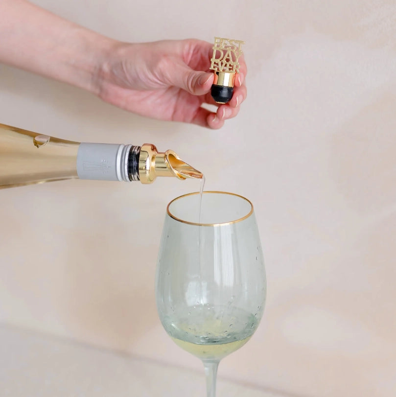 Digital photograph of a person pouring champagne from a bottle with a gold aerator into a gold-rimmed tinted glass while holding a gold Best Day Ever bottle stopper against a minimalist background