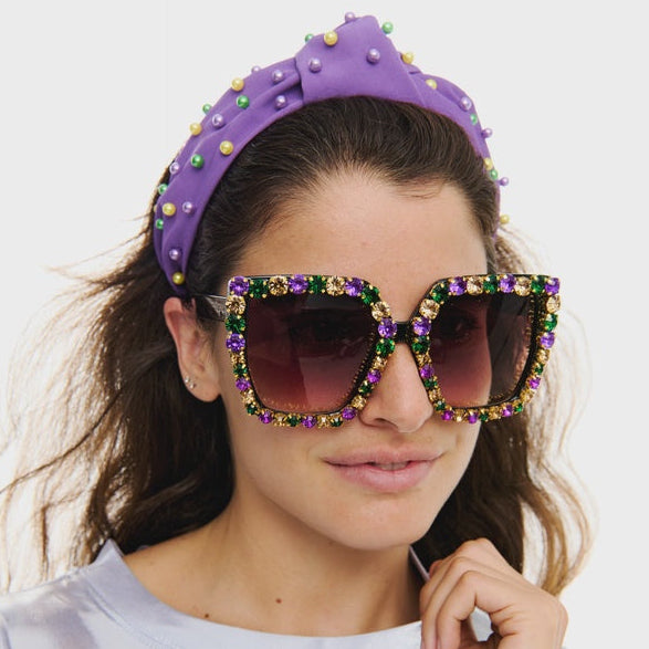Studio photograph of a woman wearing a purple knotted headband with colorful beads and oversized square sunglasses embellished with purple green and gold rhinestones in a studio setting