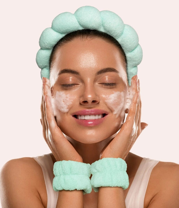 Studio photograph of a woman with glowing skin washing her face in a studio setting wearing a mint green puffy headband and matching wristbands with white cleansing foam on her cheeks