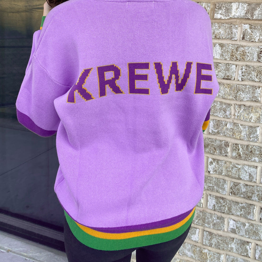 Knit sweater by KREWE worn by a person standing outdoors against a brick wall featuring iconic New Orleans Mardi Gras colors including lavender fabric, yellow-outlined purple block lettering, and vibrant green and gold striped trim
