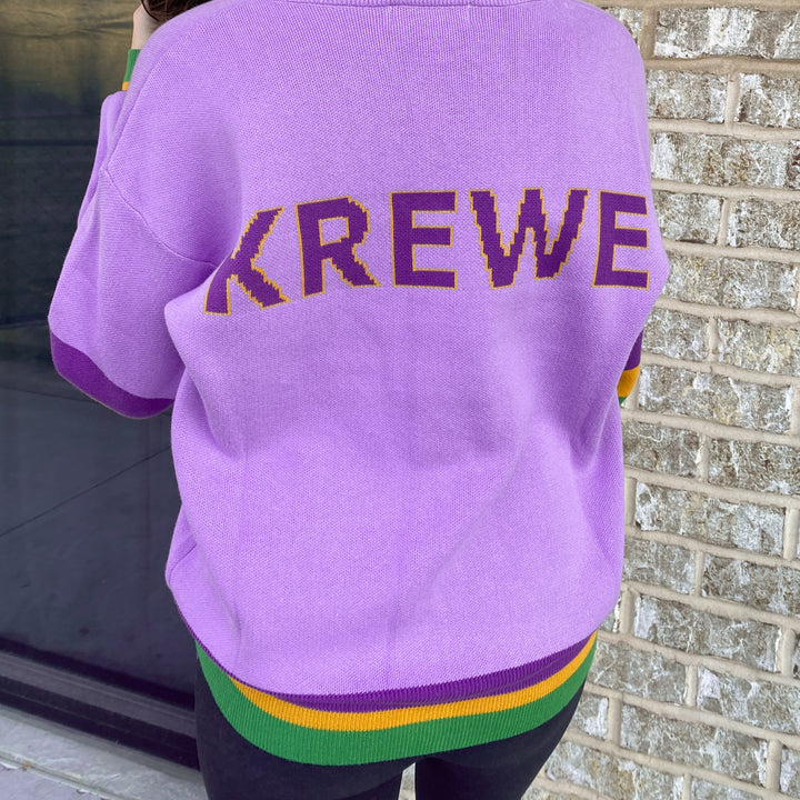 Knit sweater by KREWE worn by a person standing outdoors against a brick wall featuring iconic New Orleans Mardi Gras colors including lavender fabric, yellow-outlined purple block lettering, and vibrant green and gold striped trim