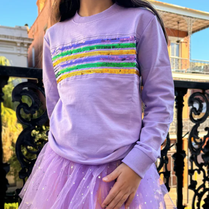 Photograph of a person in a Mardi Gras outfit on a New Orleans French Quarter balcony featuring a lavender sweatshirt with purple gold and green sequins and a purple tulle tutu with silver stars against iconic black wrought iron railings