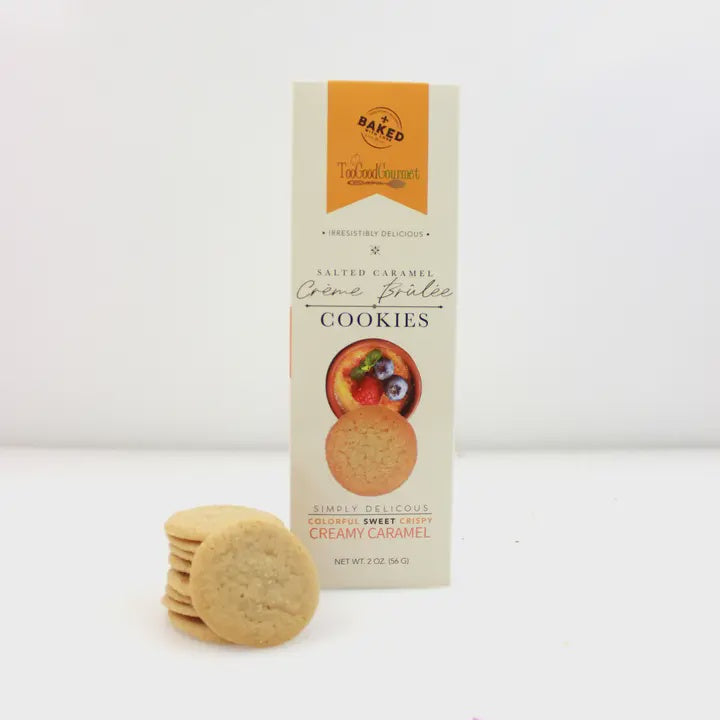 Studio photograph by Too Good Gourmet of Salted Caramel Crème Brûlée Cookies featuring a tall cream-colored box with an orange graphic and a small stack of golden-brown biscuits on a white background