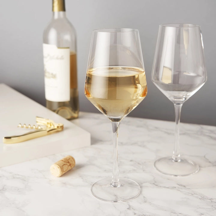 Photograph of golden white wine in a crystal glass on a marble countertop featuring a polished golden corkscrew and a wooden cork