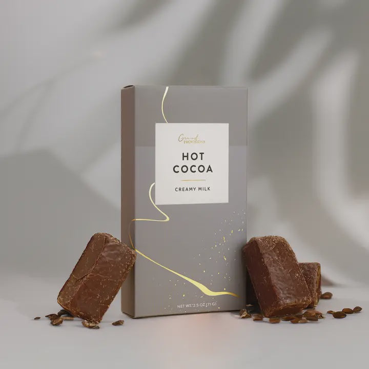 Product photograph for Grand Provisions of a Creamy Milk Hot Cocoa box in a minimalist studio setting showcasing a flowing gold foil ribbon, thick textured chocolate blocks, and soft organic leaf shadows against a neutral gray backdrop
