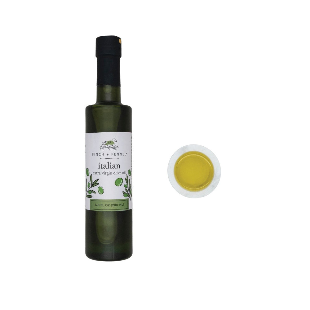 Studio photograph of Finch + Fennel Italian extra virgin olive oil in a tall dark green glass bottle with an illustrated label next to a white marble bowl of golden liquid against a white background