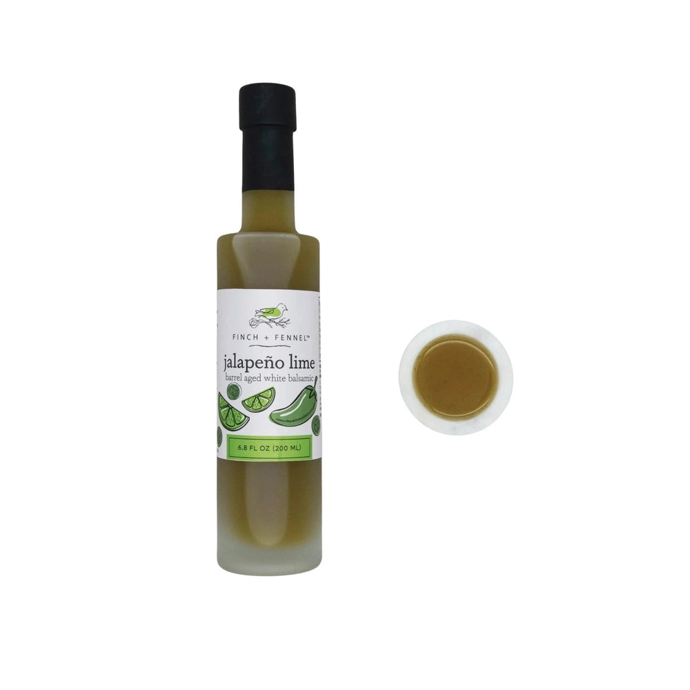 Digital product photograph by Finch + Fennel featuring a tall bottle of jalapeño lime barrel aged white balsamic vinegar with a green bird logo and a black screw cap next to a small white marble bowl of the olive-green liquid against a white background