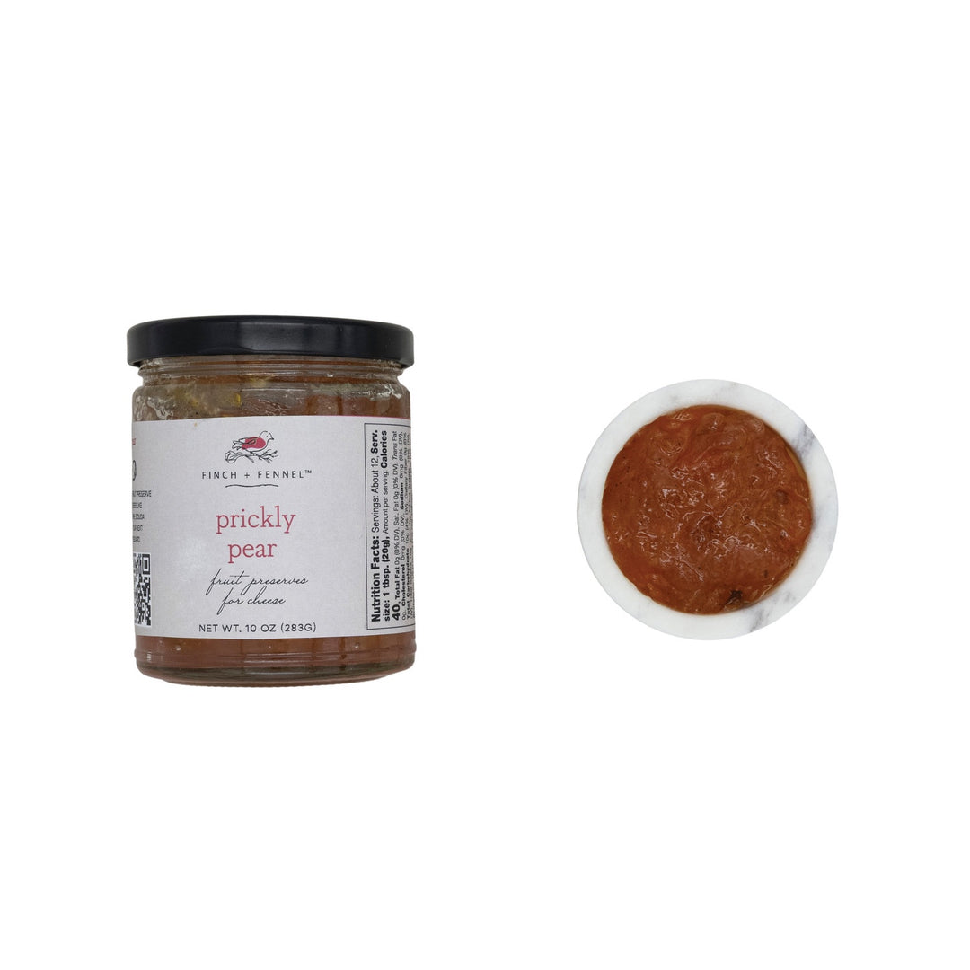 Studio product photograph by Finch + Fennel featuring prickly pear fruit preserves for cheese in a 10 oz glass jar with a black lid and a small white marble bowl filled with thick reddish-brown jam against a plain white background
