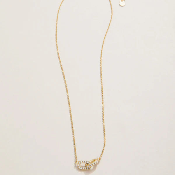 Gold necklace featuring a delicate chain and a double-link pendant with one shimmering crystal-encrusted oval link interlocking with a polished gold link on a neutral surface