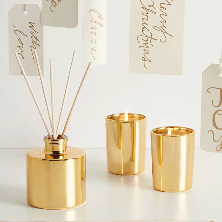 Professional product photograph of a gold home fragrance set on a white tabletop featuring a reflective metallic reed diffuser and two lit votive candles positioned before hanging holiday tags with gold calligraphy