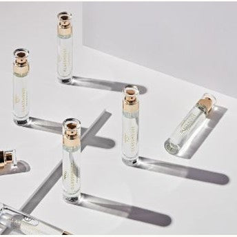 Studio photography of Glasshouse Fragrances travel perfume vials on a white minimalist surface featuring gold-toned pump dispensers, clear glass bodies, and long geometric shadows