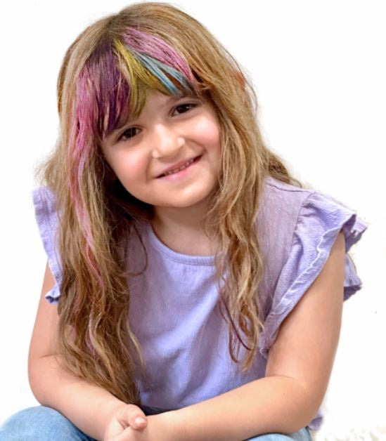 Photograph of a smiling young girl against a white background featuring long brown hair with vibrant rainbow-colored bangs and a light purple ruffled shirt