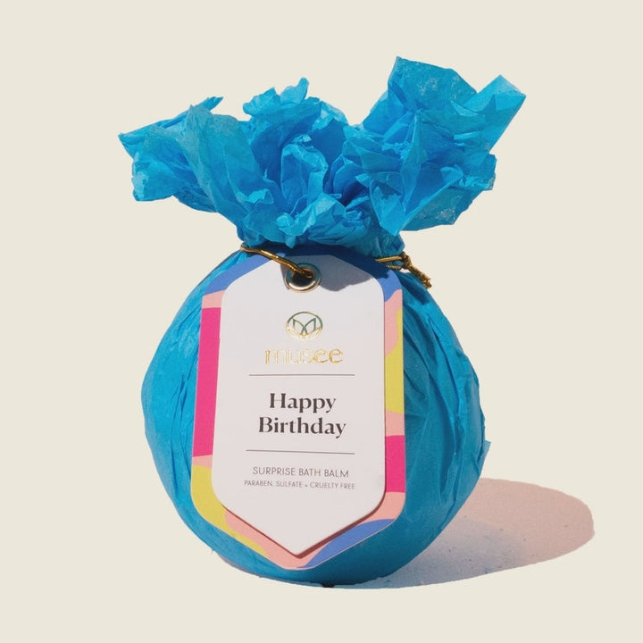 Commercial product photography by Musee of a Happy Birthday Surprise Bath Balm wrapped in vibrant blue crinkled tissue paper featuring a colorful abstract tag with a gold logo on a neutral background
