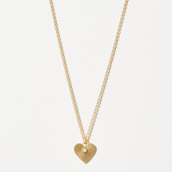 Gold jewelry necklace showcasing a heart-shaped pendant with a radiating sunburst pattern and a small central iridescent stone on a delicate cable chain against a plain white background