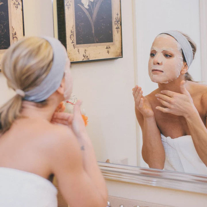 Photograph of a woman in a bathroom wearing a white sheet mask and grey headband reflected in a mirror with framed botanical prints and a white spa towel visible