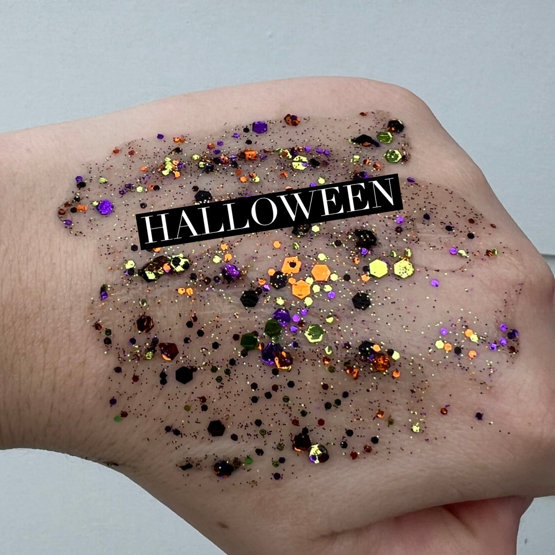 Cosmetic glitter swatch on the back of a hand featuring chunky purple and orange hexagonal glitters with a central black text banner reading HALLOWEEN