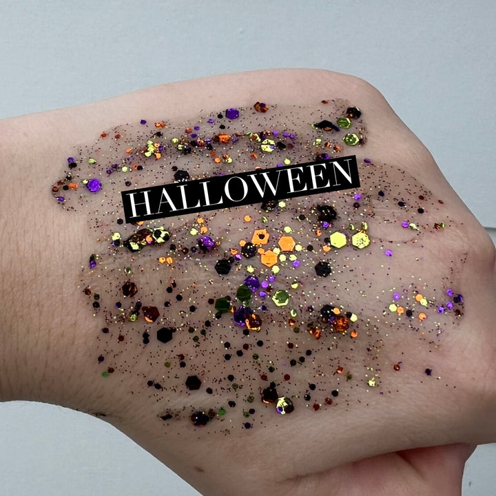 Cosmetic glitter swatch on the back of a hand featuring chunky purple and orange hexagonal glitters with a central black text banner reading HALLOWEEN