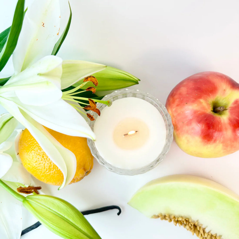 Photograph of a still life flat lay on a white surface featuring a lit candle in a fluted glass holder surrounded by white lilies with bright orange anthers, a red-and-yellow mottled apple, and a slice of green melon with textured seeds