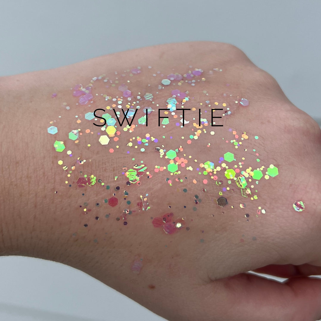Digital photograph of iridescent body glitter applied to the back of a hand featuring the text SWIFTIE surrounded by multi-sized hexagonal flakes and shimmering holographic highlights in pastel pink and lime green